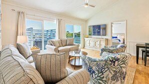A spacious living room with a sofa set, a coffee table, and a dining table. There is a large window with a view of a building and greenery outside. - Duck, Duck, Goose | 650 ft to the beach | Hot Tub (Duck)