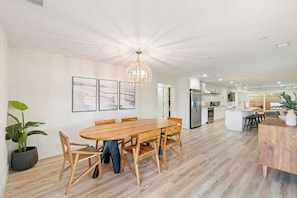 Un comedor amplio con una mesa y sillas de madera, una lámpara colgante moderna y una cocina al fondo.