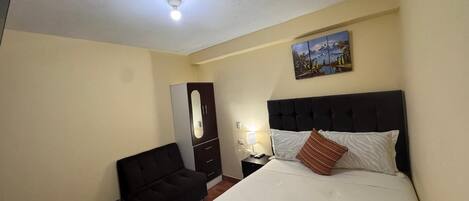 Un dormitorio con cama, mesita de noche, cómoda, una silla y un cuadro en la pared.