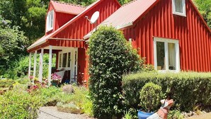 A red wooden house with a porch, surrounded by lush greenery.
