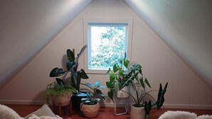 An attic room with a sloped ceiling, a window with a view of trees, and several potted plants.