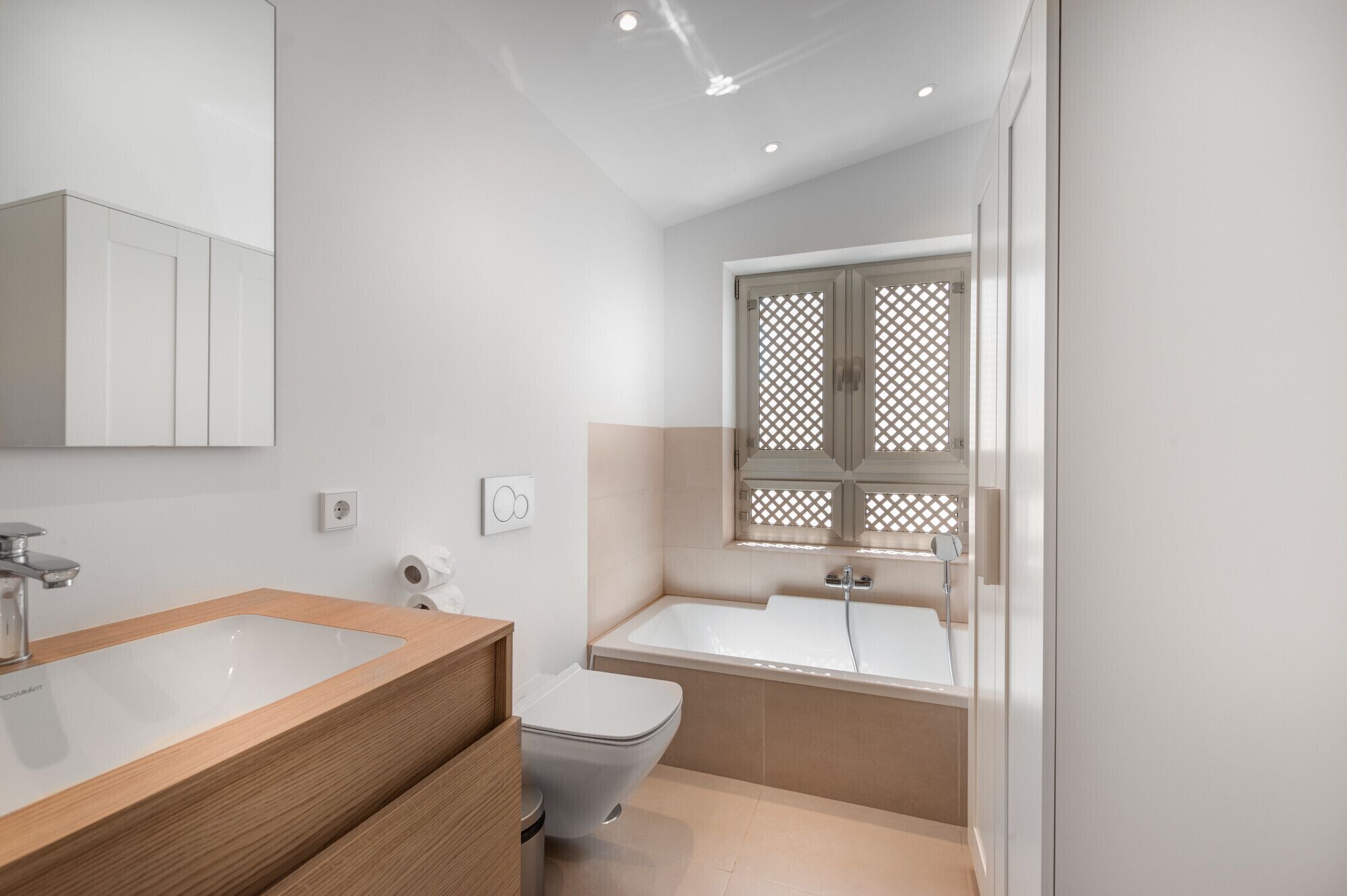 A modern bathroom with a wooden vanity, a white toilet, a bathtub, and a window with a lattice design.