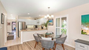 A modern kitchen with white cabinets, a stainless steel refrigerator, and a dining area with a glass table and wicker chairs. - Charming 1-bedroom condo in lovely Kihei with WiFi, AC (Kihei)