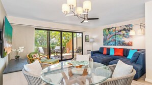 A modern living room with a glass-top coffee table, wicker chairs, a blue sofa with colorful cushions, and a large window offering a view of the outdoors. - Charming 1-bedroom condo in lovely Kihei with WiFi, AC (Kihei)