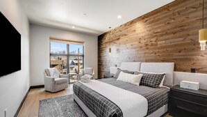 Moderni makuuhuone, jossa on suuri sänky, puusta tehty korosteseinä, näkymä ikkunasta sekä oleskelualue, jossa on tuoli ja pöytä.