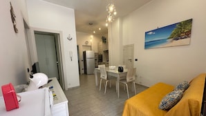 Ein modernes Wohnzimmer mit einem gelben Sofa, ein Essbereich mit weißem Tisch und Stühlen, eine Küche mit Kühlschrank und ein Flur, der zum Schlafzimmer führt.