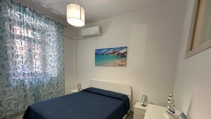 Ein Schlafzimmer mit einem Bett, einem Nachttisch, einem Schreibtisch, einer Lampe und einem Bild mit einer Strandansicht.