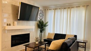 A modern living room with a flat-screen TV mounted above a fireplace, a black sofa with cushions, a coffee table, and a decorative mirror. - Luxury 3 Bedroom Home | Fireplace | 84” Pool Table| Air Conditioning| Garage (Edmonton)