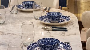 A dining table set with blue and white patterned plates, bowls, and cutlery, surrounded by modern chairs.