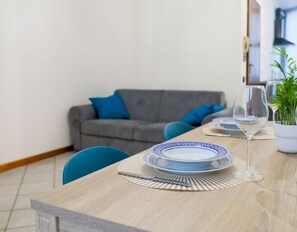 Een eetgedeelte met een tafel gedekt voor twee personen, een grijze bank met blauwe kussens, en een potplant op de achtergrond.