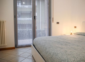 Een slaapkamer met een bed, een radiator, een schuifpui naar een balkon en een nachtkastje met een lamp.