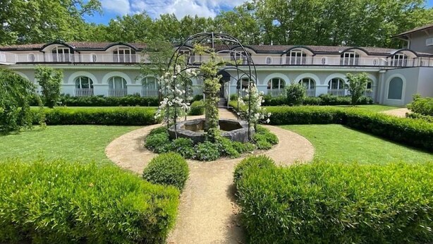 A formal garden with a central fountain, surrounded by manicured hedges and a building with arched windows in the background.
