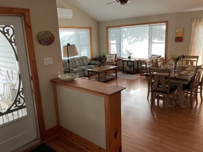 A spacious living area with a wooden dining table, a sofa, and a coffee table. There are large windows providing natural light and a view of the outdoors. - Four Season Retreat! Spacious 3-bedroom cottage on Desert Lake with WiFi, AC (Desert Lake)
