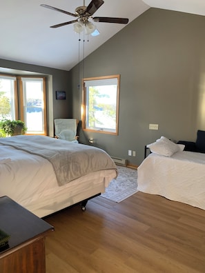 Une chambre avec un grand lit, un plus petit lit, une chaise et un ventilateur de plafond. Il y a des fenêtres et une vue sur l’extérieur.