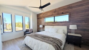 A bedroom with a large bed, two bedside tables with lamps, a ceiling fan, and large windows offering a view of the outdoors. - Red Wolf Lodge - new detached home - private hot tub - views - EV charger (Fraser)