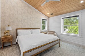 Ein Schlafzimmer mit Bett, Nachttisch, Lampe, Stuhl und Fenstern mit Blick auf Bäume.