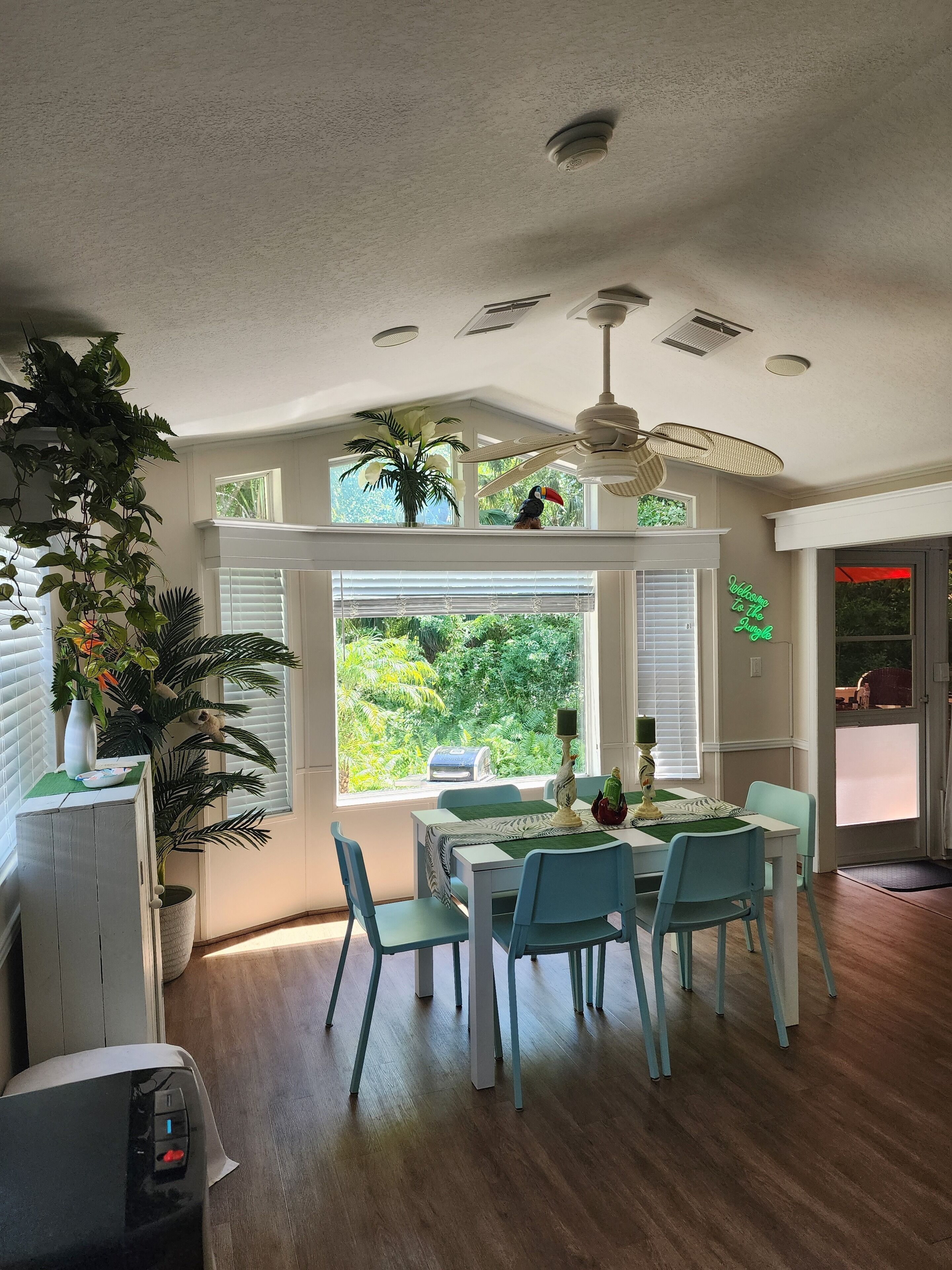 A dining area with a table set for four, surrounded by blue chairs, a ceiling fan, and a view of lush greenery through large windows.