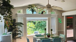 A dining area with a table set for four, surrounded by blue chairs, a ceiling fan, and a view of lush greenery through large windows. - Sun Outdoors: Clean, Private, Cozy & Loaded with Perks! Golf Cart Included (Sarasota)