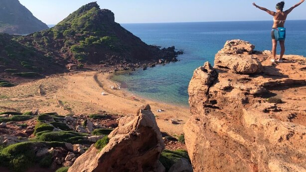A person standing on a rocky outcrop overlooking a beach with clear waters and a mountainous backdrop. - Starbal (Es Castell)