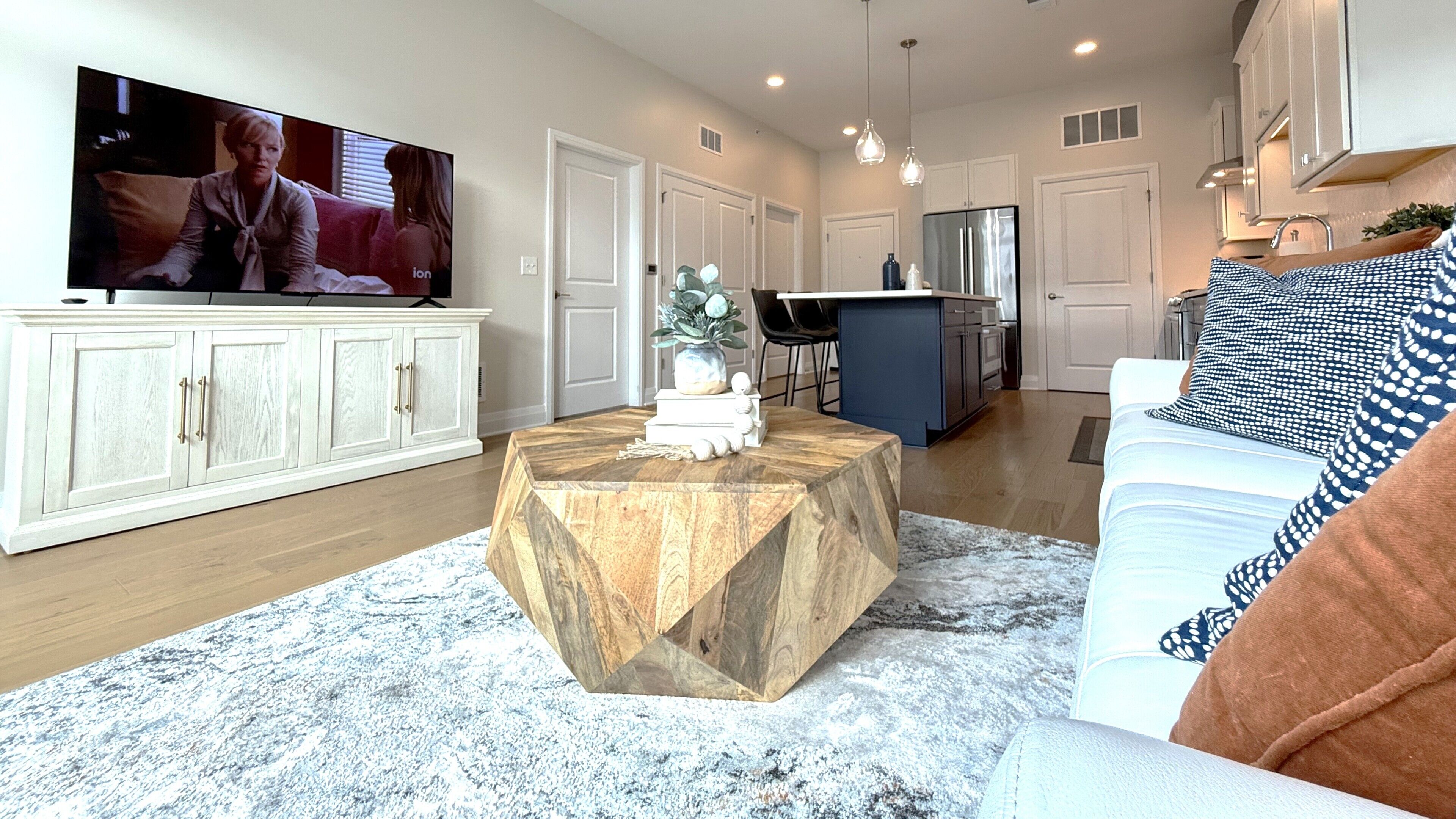 A modern living room with a large flat-screen TV, a wooden coffee table, a white sofa, and a kitchen area in the background.