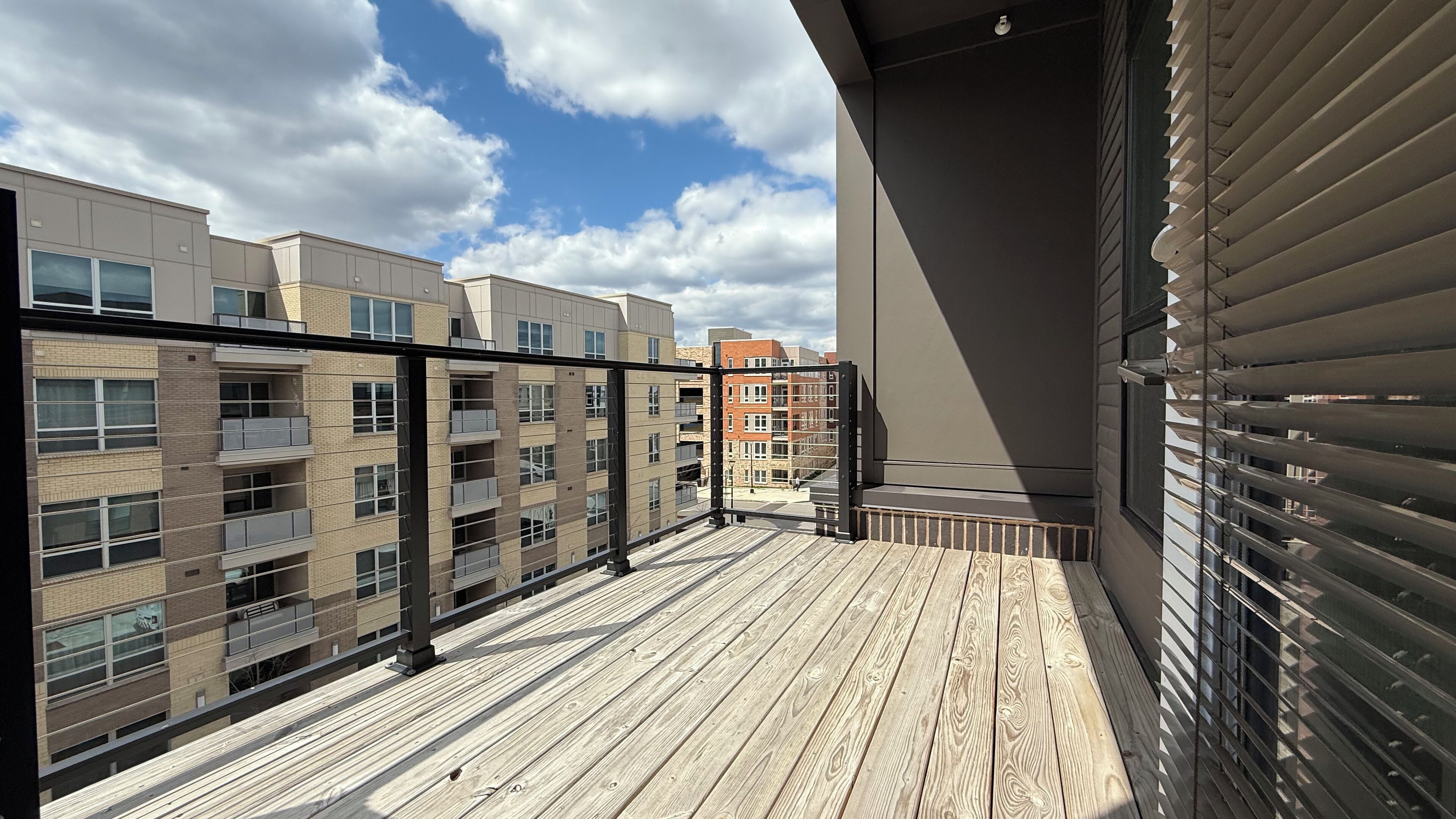 A balcony with a wooden floor and metal railings, overlooking a row of multi-story buildings with balconies.