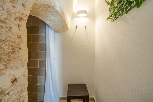 A narrow hallway with a stone archway, a small wooden stool, a wall-mounted light, and a green plant decoration.