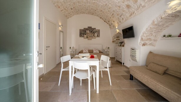 A room with a stone ceiling, a bed, a TV, a sofa, and a dining area with chairs and a table. - Velvet Home: charming studio in the heart of center Ostuni (Ostuni)