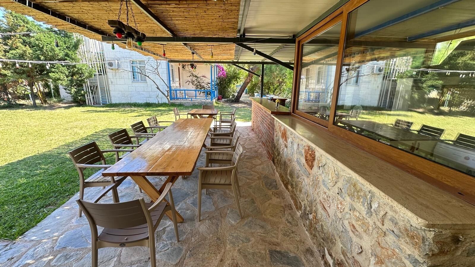 Eine Terrasse im Freien mit einem langen Holztisch und Stühlen, Steinwänden und Blick auf einen Garten.