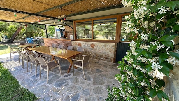 An outdoor patio with a stone floor, a wooden table, and chairs. - Zeytin Pansiyon Palamutbükü (Datca)