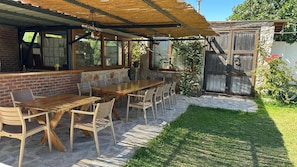 Un área de comedor al aire libre techada, con una larga mesa de madera y sillas, un muro de ladrillo y un muro de piedra con una puerta de madera.