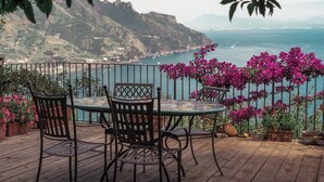 Une terrasse en bois avec une table et des chaises, offrant une vue sur un paysage côtier avec des montagnes et la mer.