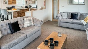 Een woonkamer met twee grijze banken, een salontafel met vier zwarte mokken, en een keuken op de achtergrond.