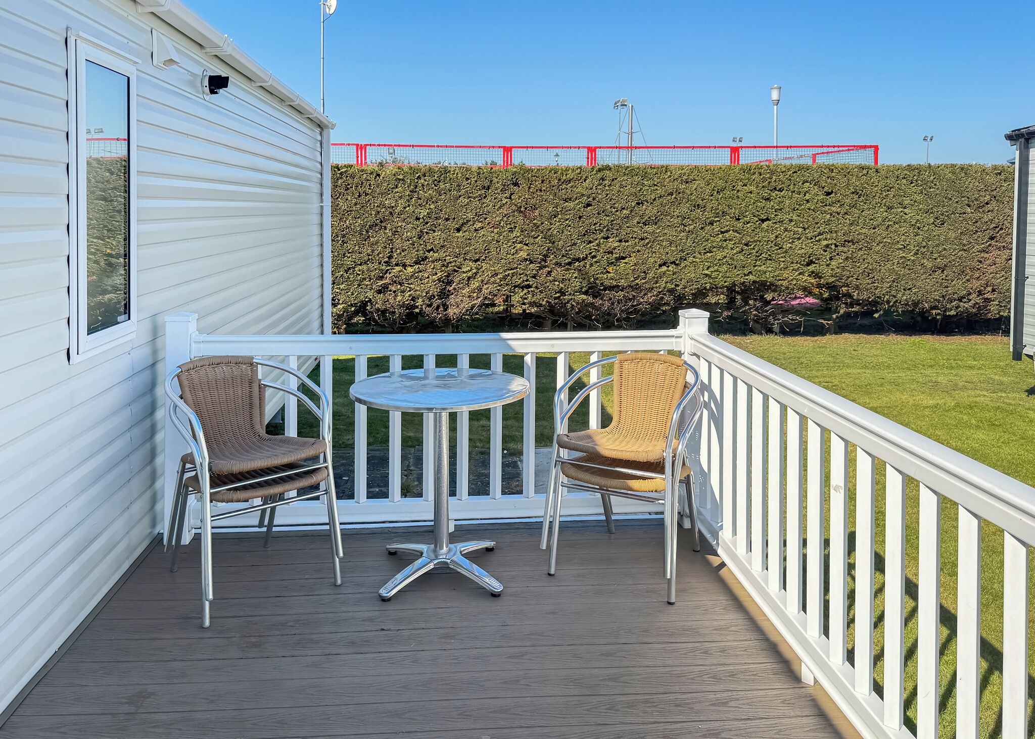 Un balcon avec deux fauteuils en osier et une petite table ronde, une balustrade blanche et une haie en arrière-plan.