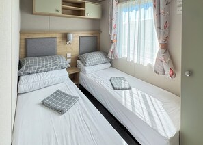 Une chambre de taille modeste, équipée de deux lits simples, d’une tête de lit avec espace de rangement, d’une table de chevet et d’une fenêtre avec des rideaux.