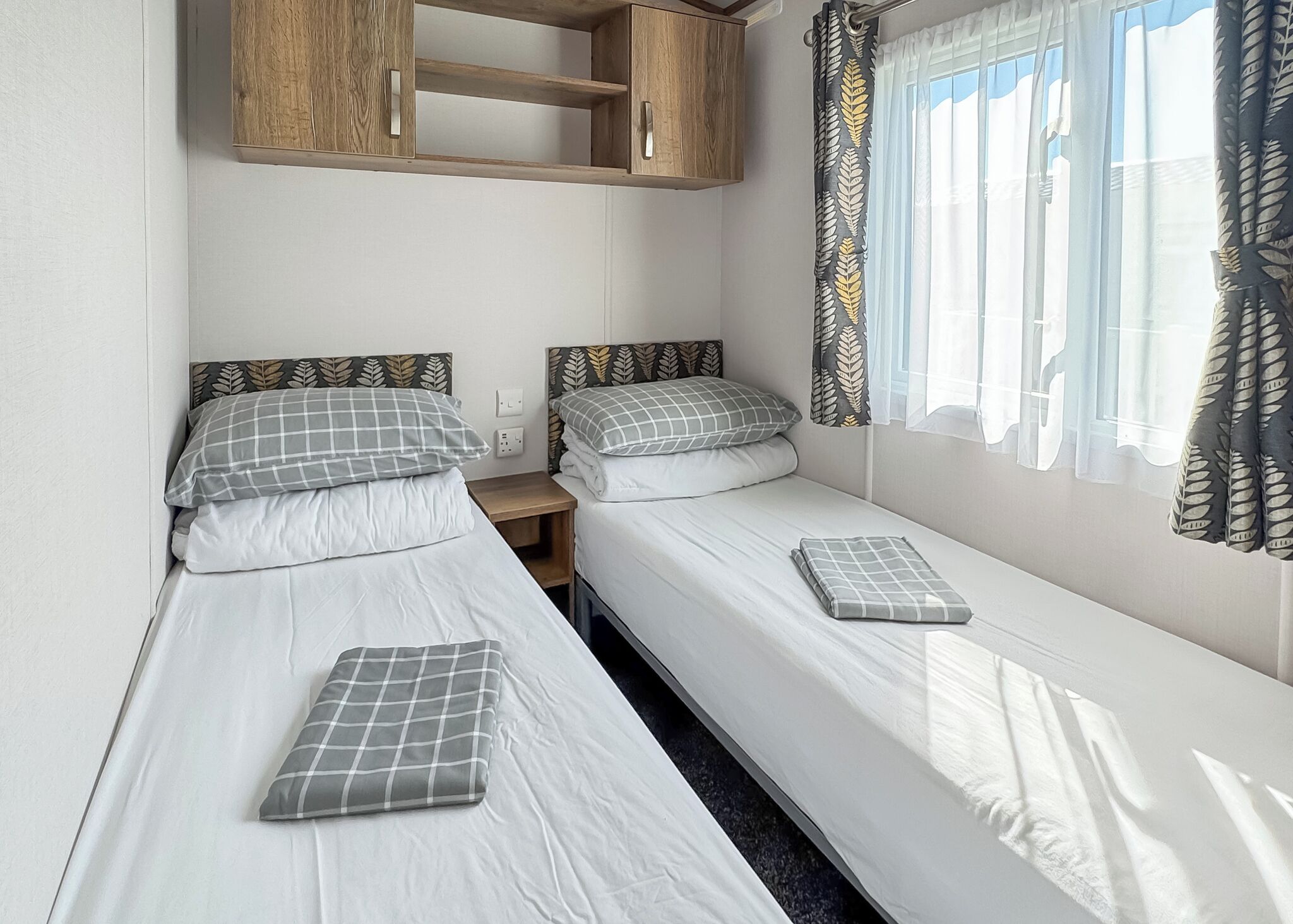 Quarto duplo com duas camas de solteiro, um armário de madeira acima e uma janela com cortinas estampadas com folhas.
