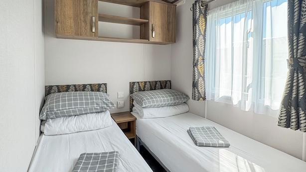 A twin bedroom with two single beds, a wooden cabinet above, and a window with leaf-patterned curtains.