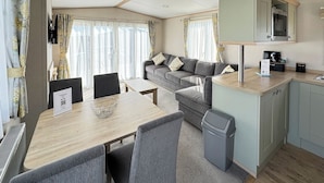 A compact, modern room with a dining table, grey upholstered sofa, and a kitchenette area.