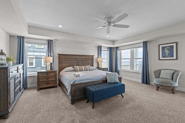 Une chambre à coucher avec un grand lit, un ventilateur de plafond, une commode, une chaise et une fenêtre avec des rideaux.