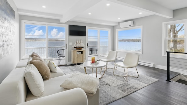 A modern living room with a large window overlooking a body of water, a white sofa, a small round table, and a chair. - Canandaigua Lakefront | Dock | Fire Pit | Kayaks (Rushville)