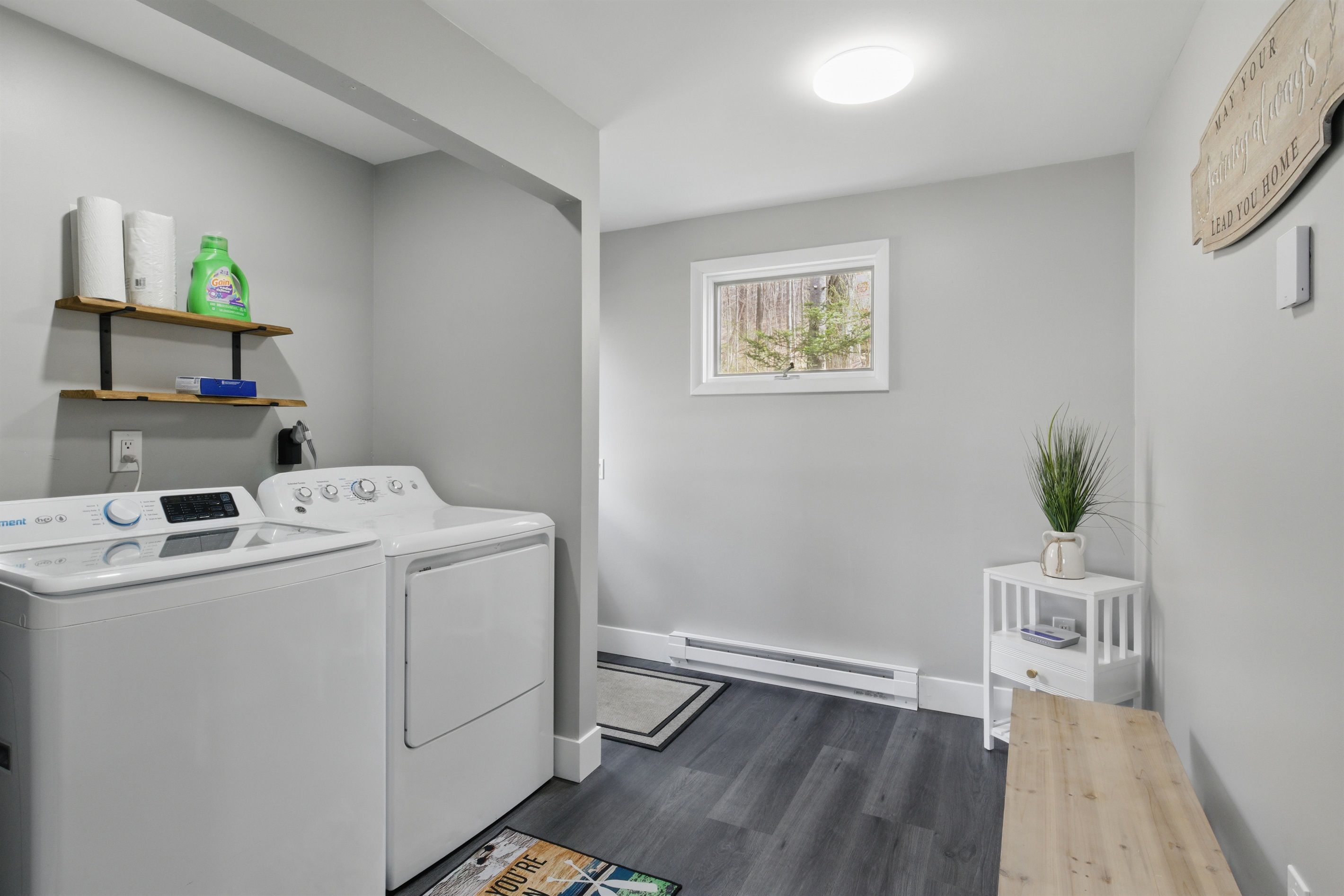 A laundry room with a washing machine, a dryer, shelves with laundry supplies, a small table with a plant, and a window with a view of trees.