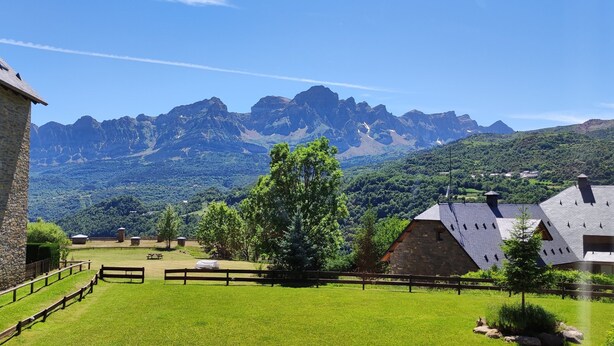 A stone building with a sloped roof, surrounded by a grassy area with trees and a view of mountains.