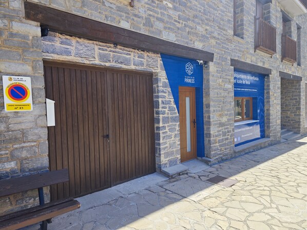 A stone building with a closed wooden door, a blue awning with a logo, and a sign indicating a permanent no parking zone.