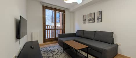 A modern living room with a grey sofa, a wooden coffee table, a TV mounted on the wall, and a large window with a view of buildings.