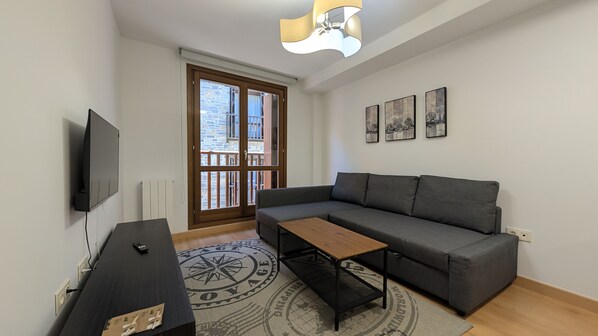 A modern living room with a grey sofa, a wooden coffee table, a TV mounted on the wall, and a large window with a view of buildings.