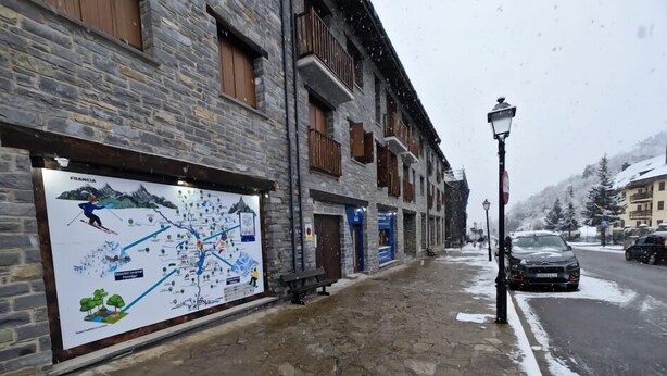 A stone-paved street with a signboard, buildings with wooden shutters, and parked cars.