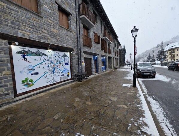 A stone-paved street with a signboard, buildings with wooden shutters, and parked cars.