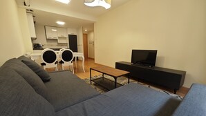A living room with a sofa, a coffee table, a TV, and a kitchen in the background.