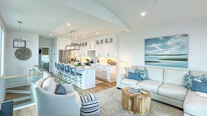 A modern living room with a sofa, coffee table, and chairs, a kitchen with white cabinets and a dining area with chairs.