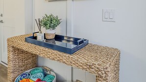 A room with a woven console table, two woven baskets, a framed painting of sailboats, and a potted plant.