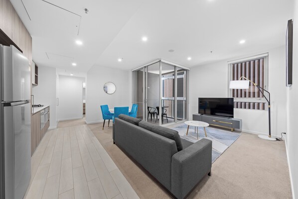 A modern living room with a grey sofa, blue chairs, a TV, and a kitchen area with a fridge. - Art House Apartments by CLLIX (South Brisbane)